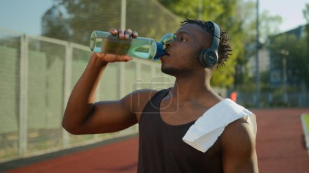 Photo for Tired African American sport man male muscular sportsman runner athlete drinking water sports bottle refresh body hydration after running on stadium workout outdoors drink listen music in headphones - Royalty Free Image