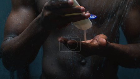 Téléchargez les photos : Afro-américain sexy nu gars lavage avec gel lotion appliquer bouteille de shampooing dans la douche sombre salle de bain athlétique nu humide homme nettoyage musculaire corps liquide savon mousse eau chaude mâle beauté bain cosmétiques - en image libre de droit