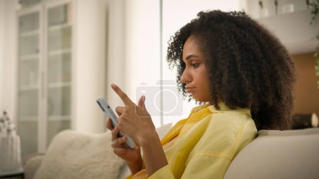 Photo for Bored African American ethnic woman gadget addict girl chatting online smartphone overuse social media addiction lazy sad female homeowner on cozy sofa at home mobile phone scrolling news on cellphone - Royalty Free Image