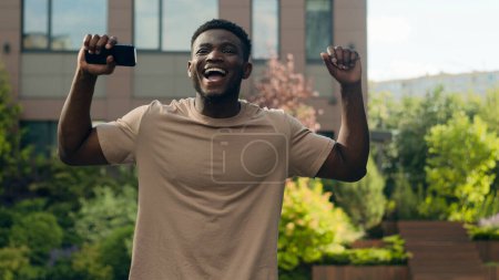 Photo for Happy excited successful joyful African American guy with smartphone success winner man with mobile phone achieve in city lucky male outdoors win victory achievement good news offer prize champion - Royalty Free Image