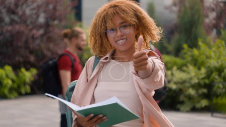 Téléchargez les photos : Afro-américaine étudiant femme lecture livre en plein air sourire à caméra montrant pouce vers le haut lycéenne écolière fille recommander comme geste sur fond de divers multiracial étudiants amis - en image libre de droit
