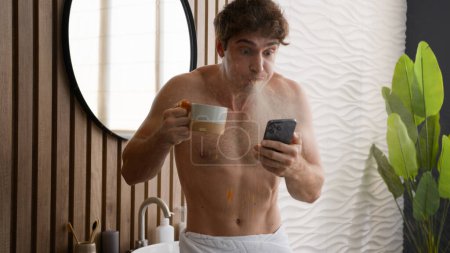 Téléchargez les photos : Caucasien homme nu regardant téléphone mobile boire du thé avec tasse dans la salle de bain torse nu gars matin hygiène vérifier les médias sociaux lire choqué nouvelles cracher boisson sentir choc confus merveille cracher café - en image libre de droit