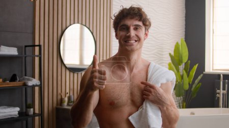 Téléchargez les photos : Nu sexy torse nu ajustement caucasien heureux homme dans salle de bain matin procédure d'hygiène bain avec serviette sur épaule souriant montrant à caméra pouce up guy mâle recommander cosmétique soins de la peau comme geste - en image libre de droit