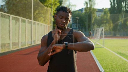 Téléchargez les photos : Homme afro-américain coureur jogger athlète sportif musclé entraînement en plein air athlétique ethnique gars en utilisant bande intelligente vérifier le pouls du cou résultat de la fréquence cardiaque sur smartwatch avant de courir sur le stade de sport - en image libre de droit