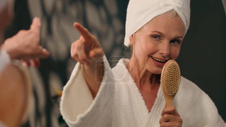 Photo for Funny mature middle-aged 50s woman in bathrobe sing at hairbrush in bathroom mirror reflection happy old female lady singing song like singer having fun dance after shower spa procedure beauty routine - Royalty Free Image