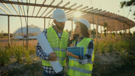 Photo for Two Caucasian professional engineers constructor man holding scheme plan woman controller check tablet talk discuss analysing ecology project outside meadow countryside. Teamwork planning engineering - Royalty Free Image