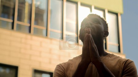 Téléchargez les photos : Concept religieux Afro-Américain priant Dieu pour l'aide prière gars suppliant bénédiction dans la ville silhouette masculine au soleil en plein air au soleil chemise soleil paumes dans la prière foi espoir souhait pardon - en image libre de droit