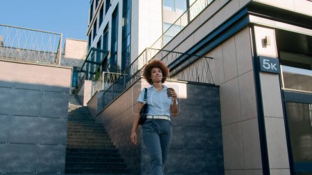 Téléchargez les photos : Plan mouvant heureux confiant afro-américain femme d'affaires dame ethnique femme d'affaires marche dans la ville marche aller déplacer tenant tasse emporter café thé boire immeuble urbain bureau skyline - en image libre de droit
