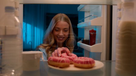 Point of view POV from inside refrigerator hungry Caucasian woman girl at night evening kitchen open fridge take out eating sweet donut unhealthy nutrition obesity diet failure overeating food addict