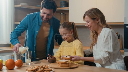 Foto de Caucasian family cooking breakfast together at kitchen at home mother with little daughter child girl prepare toasts bread with chocolate pasta father pour milk into glasses parents with kid cook - Imagen libre de derechos