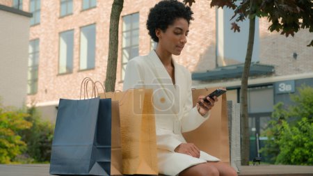 Téléchargez les photos : Élégant à la mode afro-américaine femme acheteur fille client femme en ville à l'extérieur détente après avoir magasiné téléphone téléphone téléphone téléphone achat sacs rabais ventes noir vendredi - en image libre de droit
