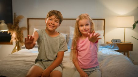 Téléchargez les photos : Deux petits enfants caucasiens sympathiques enfant enfants garçon fille frère soeur regardant caméra saluant dans la chambre à coucher sur le lit vagues mains saluant ami ou parent communiquer par appel vidéo à la maison - en image libre de droit