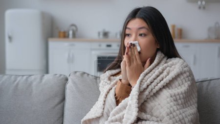 Photo for Unhealthy ill sick Asian woman multiethnic korean chinese girl patient covered in warm blanket at home blowing to paper napkin sneeze runny nose allergy flu caught cold sneezing has covid-19 symptom - Royalty Free Image