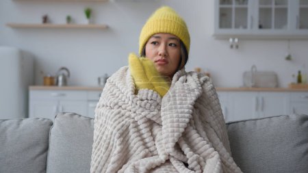 Photo for Frozen Asian chinese woman in warm hat and mittens rubbing freezing hands sit in cold kitchen frosty temperature heating problem in home apartment sick girl covered in blanket suffer from cool winter - Royalty Free Image