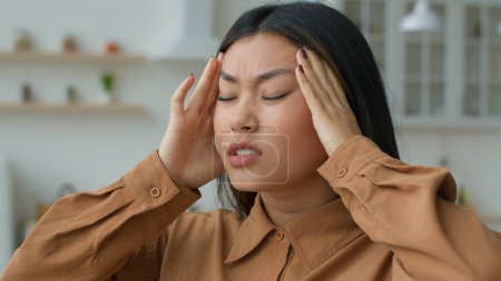 Téléchargez les photos : Fatigué malade femme asiatique se sentent mal à la tête douleur massage temples toucher mal à la tête grimace souffrir de maux migraine hypertension artérielle fille coréenne malade femme d'affaires chinoise dans la cuisine - en image libre de droit