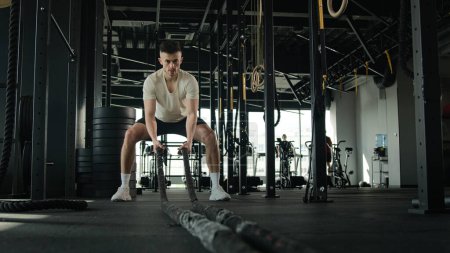 Photo for Determined Caucasian sportsman active athlete man bodybuilder training in gym muscular guy masculine workout with battle ropes sport equipment in fitness club physical exercise lifting weights effort - Royalty Free Image