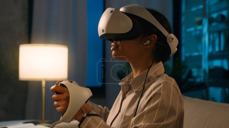 Téléchargez les photos : Femme afro-américaine casque VR lunettes réalité augmentée métaverse simulation de jeu explorer 3D monde jouer jeu vidéo technologies virtuelles modernes cyber jouer avec des contrôleurs à la maison de nuit - en image libre de droit