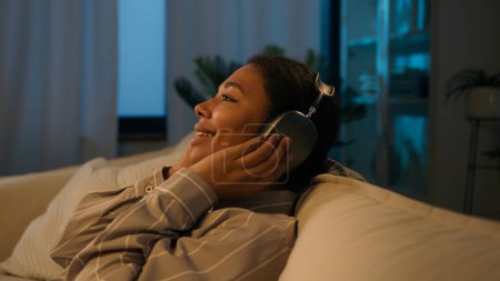 Téléchargez les photos : Femme afro-américaine détendue dans les écouteurs tombent sur le canapé confortable soirée loisirs à la maison écouter de la musique ethnique fille repos avec les yeux fermés profiter du son dans les écouteurs chanter chanson relaxante - en image libre de droit