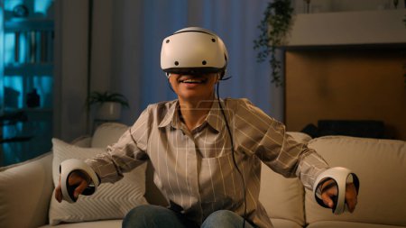 Foto de Emocionada mujer africana jugando juego de realidad virtual en casa noche feliz chica americana en la simulación de juegos VR gafas casco y controladores jugar natación 3D metaverso innovadora tecnología del ciberespacio - Imagen libre de derechos