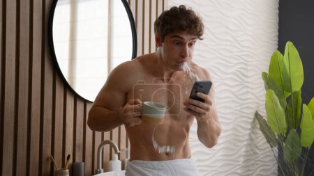 Téléchargez les photos : Caucasien détendu homme nu boire du thé à la lecture de routine du matin nouvelles dans les médias sociaux dans smartphone navigation au téléphone mobile boire du café chaud dans la salle de bain gars stupéfait choc cracher boisson - en image libre de droit