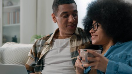 Téléchargez les photos : Heureux famille couple afro-américain mari femme avec tasse de thé détente sur canapé en utilisant ordinateur portable achats en ligne choisir des marchandises dans la boutique en ligne app discuter des offres livraison de commande ensemble - en image libre de droit