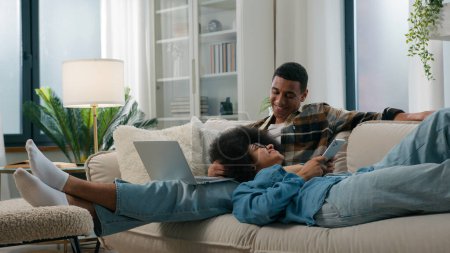 Téléchargez les photos : Frissonnant couple afro-américain détendu sur le canapé à la maison ensemble gadget internet addict homme travaillant à l'aide d'un ordinateur portable femme pose navigation téléphone mobile à l'appartement conjoints dépendance aux médias sociaux en ligne - en image libre de droit
