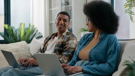 Téléchargez les photos : Femme afro-américaine travailler avec ordinateur portable homme couple de travail assis à la rangée deux pigistes avec des ordinateurs coworking à la maison famille toxicomane regardant l'autre riant jouer au jeu en ligne sur le canapé - en image libre de droit
