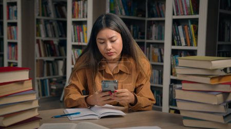 Photo for Asian girl student sitting in library write doing homework frustrated young woman getting notification on mobile phone reading bad news failing exam dropout from college losing scholarship feel upset - Royalty Free Image