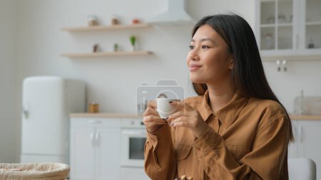 Photo for Relaxed girl Asian korean woman in domestic kitchen with cup of coffee drinking hot herbal tea multiethnic lady thoughtful calm housewife drink beverage looking in distance dreaming with mug in hands - Royalty Free Image