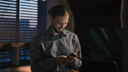 Carefree positive browsing phone mobile app Caucasian businessman at evening office using smartphone smiling happy business man entrepreneur scrolling cellphone check email work break message network