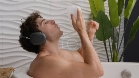 Téléchargez les photos : Calme tranquille Caucasien nu détendu gars assis dans la salle de bain homme au repos se détendre dans une baignoire mousseuse souffler mousse profiter de l'audio dans les écouteurs mâle détente à la maison bain hôtel écouter de la musique chanson dans les écouteurs - en image libre de droit