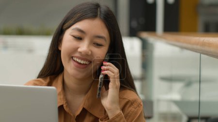 Foto de Mujer asiática sonriente empresaria que trabaja en línea con el ordenador portátil en la oficina chica japonesa china estudiante freelancer hablar teléfono móvil riendo emocionado respuesta femenina teléfono inteligente llamada conversación despreocupada - Imagen libre de derechos