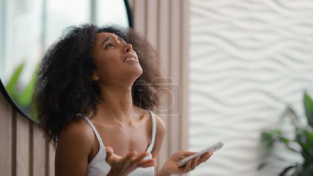 Téléchargez les photos : Afro-Américain mécontent jeune femme dans la salle de bain à la maison baignoire en regardant l'écran du smartphone insatisfait de mauvaises nouvelles message spam escroquerie sms ethnique biracial fille malheureux souligné panne de téléphone mobile - en image libre de droit