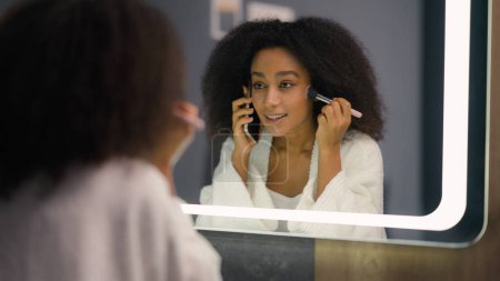 Female mirror reflection happy girl talking phone mobile call chatting beauty visage make-up biracial beautiful African American woman apply powder for face tone preparing cosmetics talk smartphone