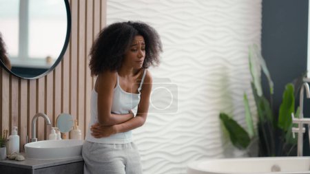 Téléchargez les photos : Afro-américaine malade malade malaise triste femme avec maux d'estomac debout dans la salle de bain à la maison dans le bain fille ethnique biraciale souffrant de douleurs abdominales règles douloureuses digestion problème de santé - en image libre de droit