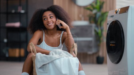 African American smiling happy woman biracial ethnic female girl lady housewife housekeeping sit on floor talking mobile phone with basket of laundry near washing machine talk smartphone chatting