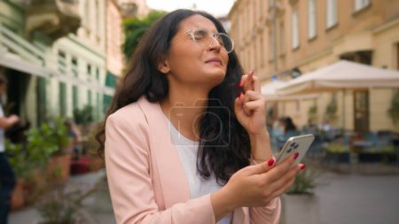 Téléchargez les photos : Heureuse Indienne Arabe femme d'affaires femme croisée doigts priant geste désirant regarder entreprise de téléphonie mobile en dehors de la ville rue café femme gagnant célébrant victoire espoir chance triomphe se réjouir - en image libre de droit