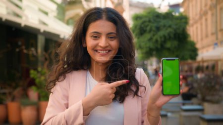 Foto de Feliz indio árabe étnica mujer de negocios mantener el teléfono móvil en la ciudad al aire libre sonriente mujer señalando el dedo en la pantalla verde croma clave copia espacio mockup smartphone para publicidad aplicación de negocios - Imagen libre de derechos