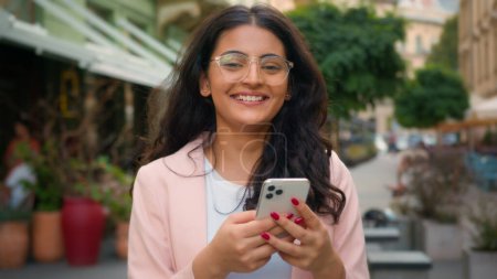 Téléchargez les photos : Heureuse femme souriante indienne Arabe ethnique femme utilisateur client tenant téléphone portable en dehors du café de rue de la ville. Client positif hochant la tête oui bonne réaction approuver la technologie gadget - en image libre de droit
