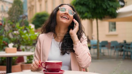 Téléchargez les photos : Gros plan joyeux excité Indien Arabe ethnique femme fille femme d'affaires voyageur parlant appel téléphonique mobile réjouissant sourire agréable conversation lointaine à l'extérieur de la ville café restaurant - en image libre de droit
