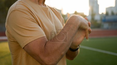 Téléchargez les photos : Vieil homme caucasien étirer les mains avant l'entraînement échauffement stade en dehors de la ville sportif sport style de vie rayons du soleil endurance activité physique bien-être motivation bodybuilder flexibilité aérobic exercice - en image libre de droit