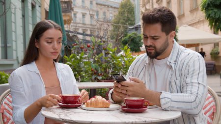 Caucasian couple eat breakfast in city cafe outdoors addicted busy man boyfriend using mobile phone ignoring upset woman girlfriend offended arms crossed quarrel crisis relationship problem conflict