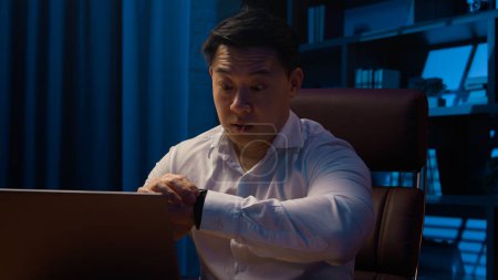 Téléchargez les photos : Adulte asiatique homme chinois travailleur coréen homme d'affaires travaillant dur ordinateur portable en ligne projet date limite tard dans le bureau à domicile fatigué à regarder montre-bracelet vérifier le temps soir surchargé mâle - en image libre de droit