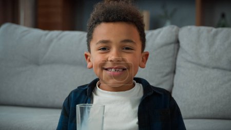Kopfschuss Nahaufnahme Porträt kleine lustige niedliche 10s Kind Junge ethnische Kind trinken Milch zu Hause lächelnd in die Kamera mit schiefen Zähnen. Afroamerikanischer Sohn trinkt Glas süßen Bio-Joghurt glückliches Lächeln