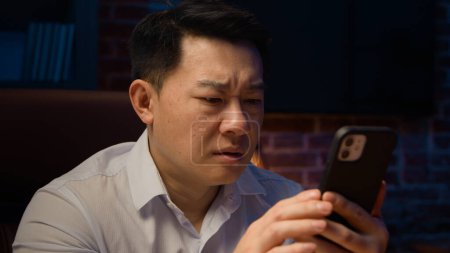 Téléchargez les photos : Asiatique ethnie 40s homme la nuit soir bureau à domicile tenant téléphone mobile se sentent malheureux émotions négatives bouleversé par recevoir de terribles mauvaises nouvelles coréen adulte homme d'affaires avec smartphone échec perdu - en image libre de droit