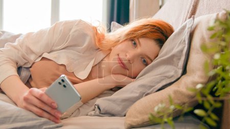 Téléchargez les photos : Gros plan Femme caucasienne avec smartphone couché au lit se détendre le matin dans la chambre à coucher maison bavarder avec téléphone mobile femme blogueuse influenceuse fille défilant les médias sociaux en ligne sourire après le réveil - en image libre de droit