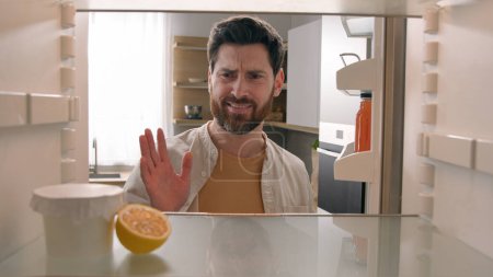 Photo for POV point of view from inside refrigerator hungry Caucasian man at kitchen open empty fridge with half of lemon and jam yogurt jar nothing to eat dissatisfied disgust close door food delivery service - Royalty Free Image