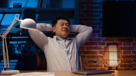 Téléchargez les photos : Productif homme d'affaires coréen Asiatique homme d'affaires chinois travailleur penché en arrière finition travail sur ordinateur portable soulagement de l'ordinateur après le travail fait étirer les mains détendues tâches terminées au bureau du soir - en image libre de droit