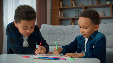 Foto de Dos chicos afroamericanos étnicos disfrutan del arte del hobby en casa, los deberes, dibujan, los escolares, los hermanos, los hermanos, los niños, dibujan con marcadores de colores, crean imágenes con plumas de fieltro de colores, pintan - Imagen libre de derechos