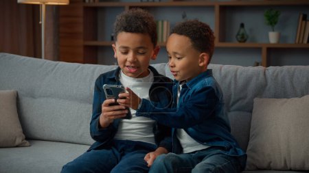 Photo for Two little African American siblings boys children kids little brothers with mobile phone smartphone playing game online talking browsing sit at couch at home with cellphone app scrolling web page - Royalty Free Image